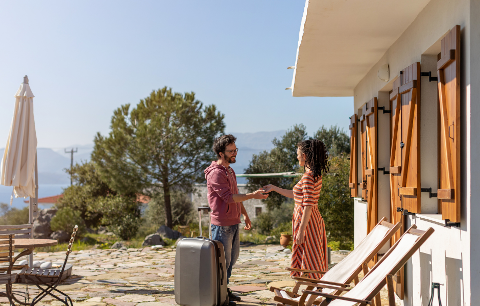 A man and woman shaking hands outside a vacation property, symbolizing a successful real estate deal. The setting includes a sunny outdoor patio with wooden chairs, greenery, and a scenic view, emphasizing financial freedom with real estate investing.