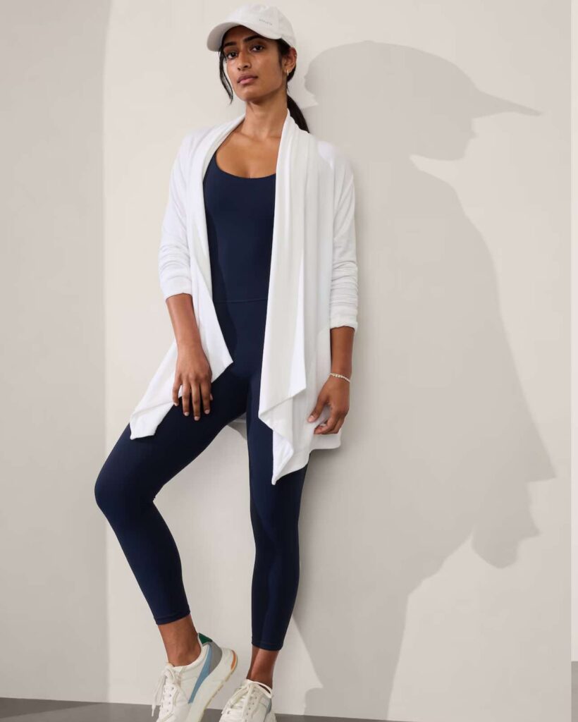 An image of a woman leaning against a wall with one leg bent and the foot placed on the wall. The woman is wearing dark leggings, a dark top, white long cardigan, and light-colored sneakers. Image used for the article Tunic and Leggings Combo.