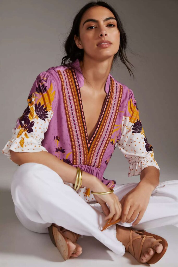 Close-up image of women seated wearing a colorful patterned top with purple and orange hues, white pants, and brown strappy sandals. The woman is also wearing multiple bracelets on both wrists, showing perfect Disney Worl mom outfits 