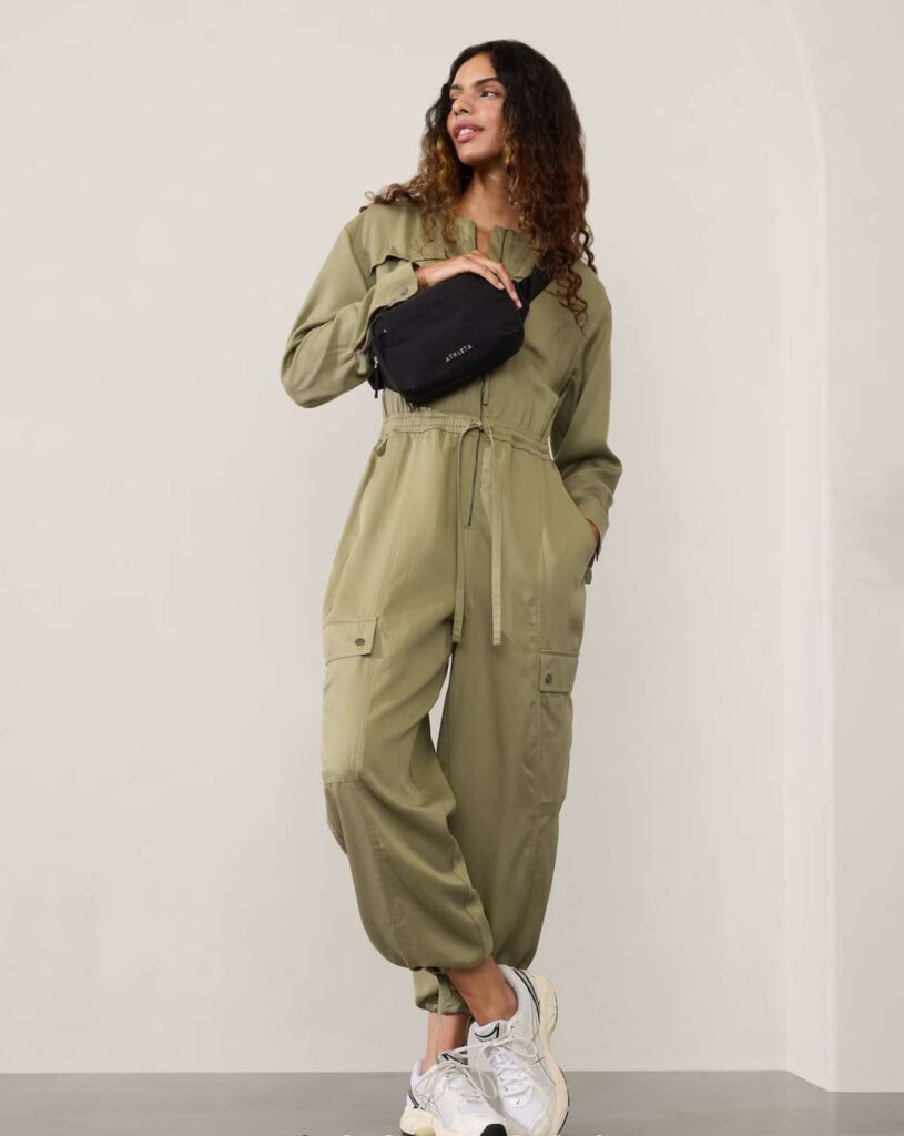 Image of a woman wearing a stylish, olive green jumpsuit stands confidently against a plain background. The jumpsuit features a belted waist and long sleeves, giving it a chic and modern look. She pairs the outfit with black ankle boots, adding a touch of elegance to her ensemble. Used for the article What to Wear to Disneyland