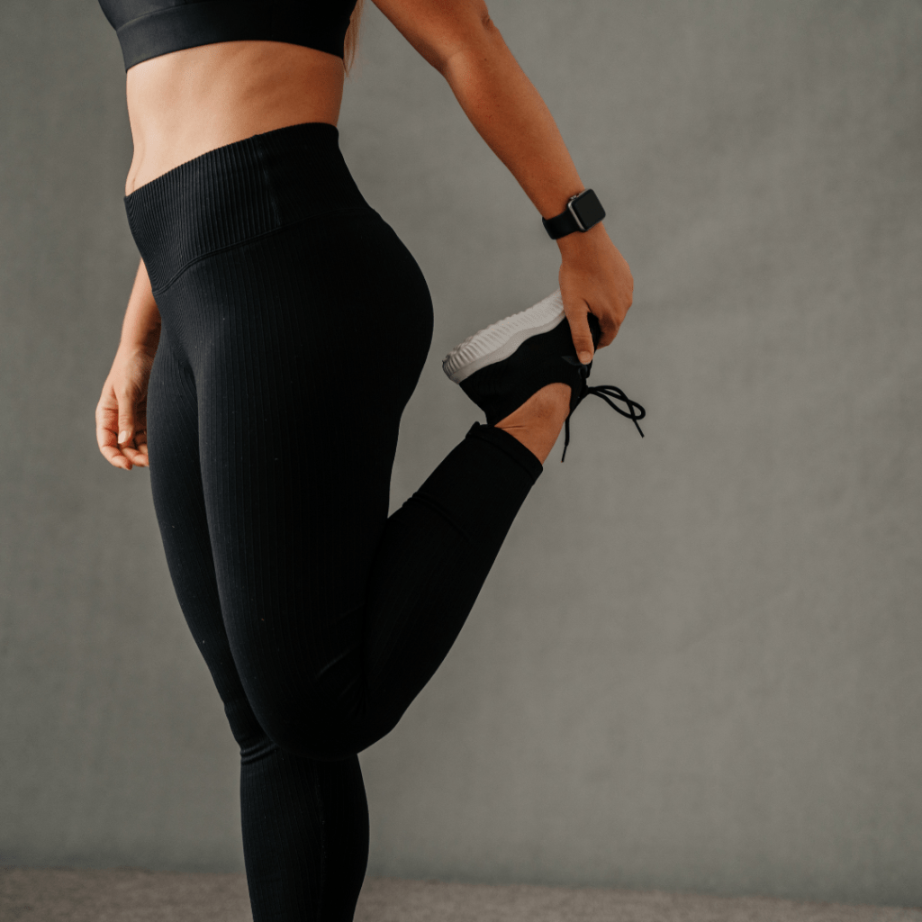 Close-up image of a person showing from the waist down, stretching their leg muscles. They are wearing black sports leggings and a black top, with a smartwatch on their left wrist. The individual is holding their right foot behind them, likely performing a quadriceps stretch. Image used for the article pregnancy outfit ideas for winters.