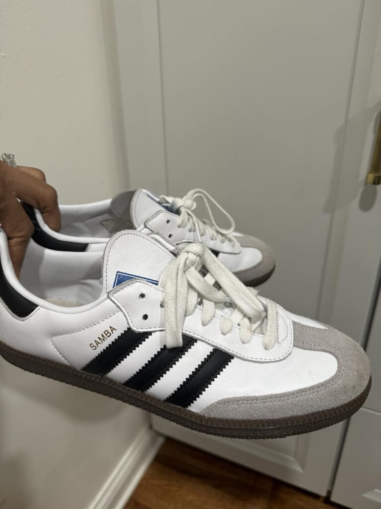 A close-up image of a pair of white Adidas Samba sneakers with black stripes, held by a person against a white door. The shoes feature the distinctive tan gum rubber sole and are laced up with white laces. The image captures the classic design of the sneakers, highlighting their condition and style. 