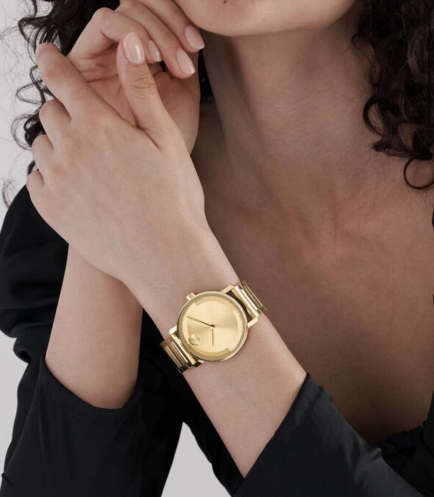 A close-up image of a person’s wrist wearing a gold watch with a simple, elegant face and no numerals. The person has manicured nails with pale polish and is wearing a black garment with a ruffled texture, which can be seen in the background. The focus is on the watch, indicating it may be an advertisement or a showcase of the accessory.