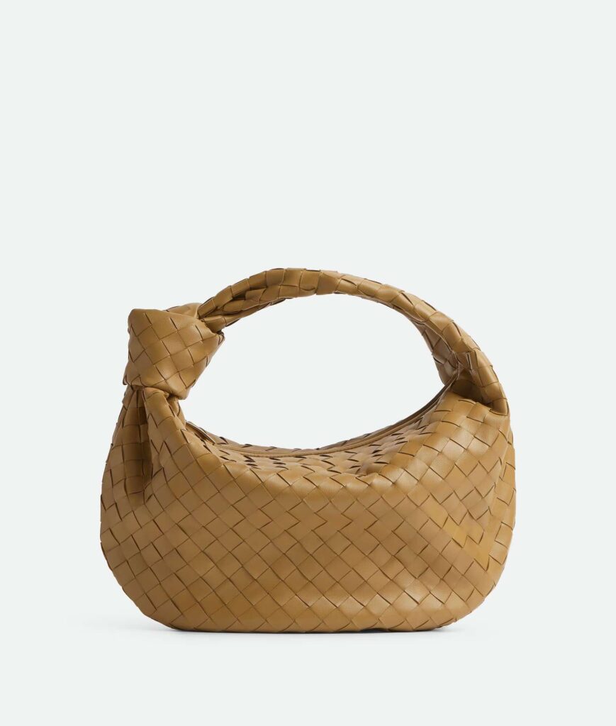 Image of a single tan-colored woven handbag with a unique crescent shape, featuring an intricate herringbone pattern. The bag has a prominent, rounded handle that seamlessly blends into the body of the bag. The background is plain and light-colored, providing a stark contrast that highlights the detailed craftsmanship of the handbag, a must-have Mom's Wardrobe Staples for 30s.