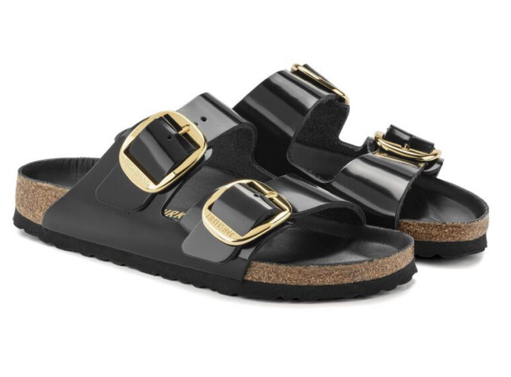 A pair of black Birkenstock sandals with two straps each, featuring large gold-toned adjustable buckles. The footbed appears to be made of cork and the sole is black. The background is a plain, light color providing contrast to the dark-colored sandals. Image used for the article A Pair of Strappy Sandals with Soft Soles.