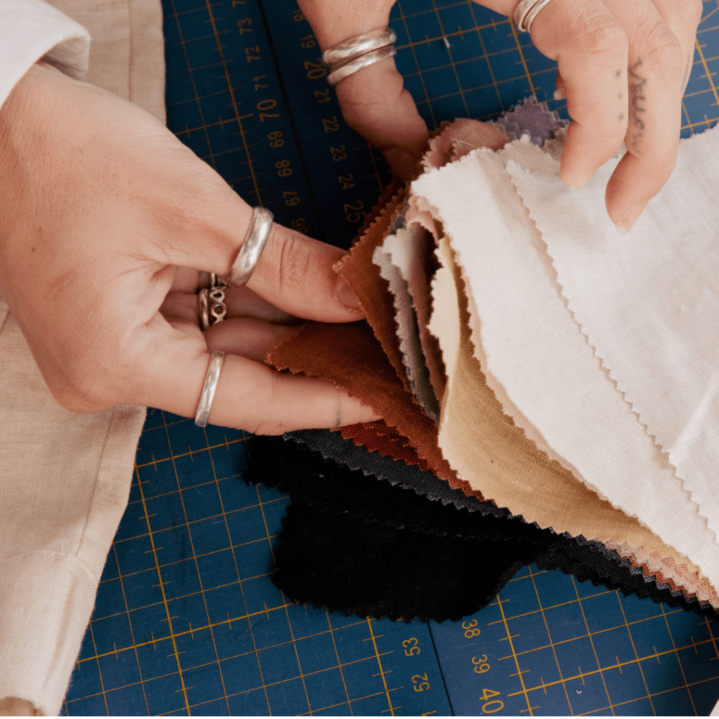 A close-up image of two hands with rings on the fingers flipping through a variety of fabric swatches with zigzag edges. The fabrics are in an assortment of colors, including white, beige, purple, red, and black. The swatches are placed on a cutting mat with measurements that indicate the mat is marked in centimeters. The hands appear to be selecting a fabric for a project, showcasing the stay at mom capsule wardrobe year-round.