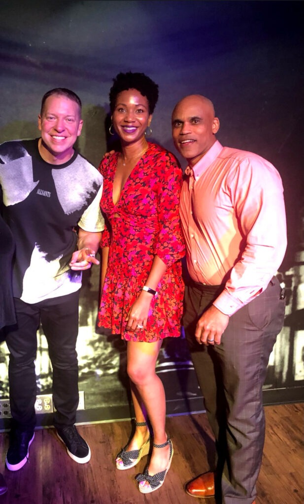 Image of three people posing together in a casual, friendly group photo. On the left is a man wearing a black and white graphic T-shirt with black pants and sneakers. In the middle is a woman dressed in a red floral-patterned wrap dress and wedge heels, accessorized with large hoop earrings. On the right is a man in an orange button-down shirt with dark trousers, smiling at the camera. They are standing on a wooden floor with a dark backdrop, and the lighting gives the scene a warm, vibrant feel showing the subtle over bold.