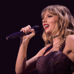 Close-up image of Taylor Swift holding a microphone close to where the mouth would be. She is wearing a shiny, sequined garment that reflects light, suggesting a performance or entertainment setting. The background is dark, highlighting the figure in the foreground. Image used for the article Taylor swift concert outfit ideas for moms.