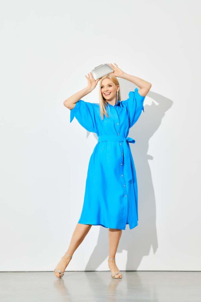 Image of a woman stands against a plain, light-colored background, wearing a bright blue dress with a belt and beige high-heeled shoes.  They have long, straight blonde hair that reaches past their shoulders, making Best Summer Fashion Outfit for Moms in Their 30s