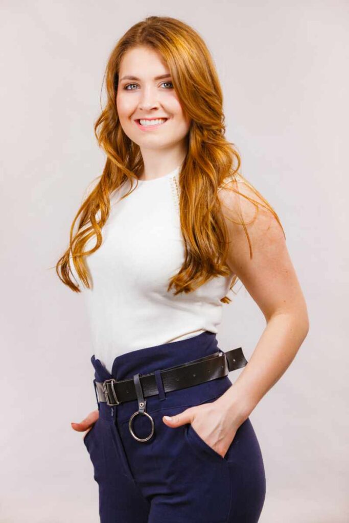 Close-up image of a woman from the waist up, facing to the side. The individual has long, wavy hair and is wearing a white sleeveless top tucked into high-waisted navy blue trousers secured with a black belt that has a large circular buckle. The background is plain and light-colored, providing a neutral backdrop that contrasts with the outfit. Best fit when no sure to what to wear in your 30s