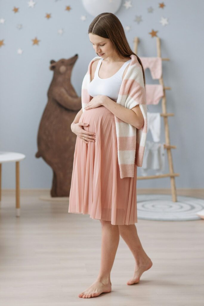 Image of a woman wearing a white top, pink skirt, and a white and pink striped cardigan stands in a room with a blurred background. They are cradling their belly with both hands, suggesting pregnancy. In the background, there is an illustration of a bear on the wall, some stars, a small table, and part of a ladder or shelving unit. Image used for the article Elegant Maternity Outfits for 12 Weeks Pregnant.