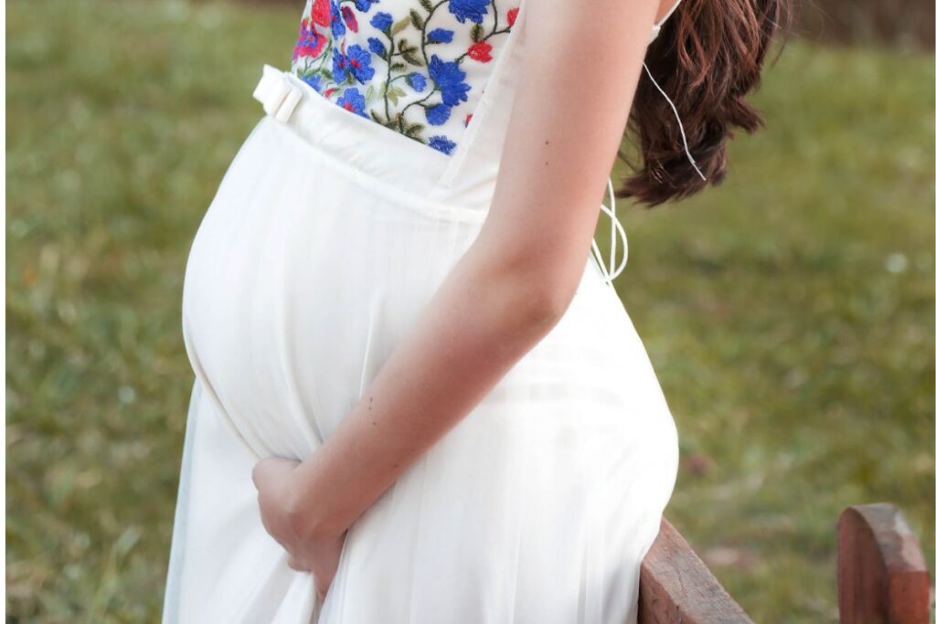 Close-up image of a pregnant standing sideways, cradling their belly with both hands. They are wearing a long, flowing dress and are outdoors in a natural setting with greenery and trees in the background. The sunlight filters through the leaves, creating a serene and peaceful atmosphere. Image used for the article What Pants to Wear During Pregnancy to Look Stylish.