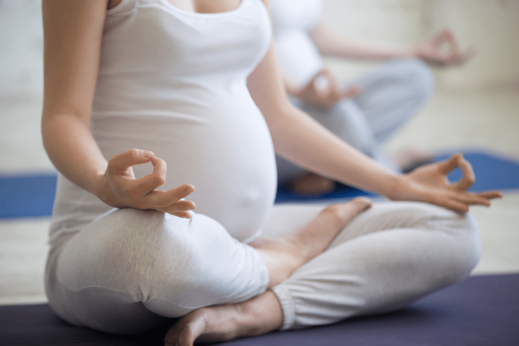 A close-up image of a pregnant individual practicing yoga in a seated position with their hands resting on their knees in a mudra gesture. They are wearing white yoga attire. In the blurred background, another individual can be seen in a similar pose, suggesting a group prenatal yoga class setting. Image used for the article Casual Winter Pregnancy Capsule Wardrobe.