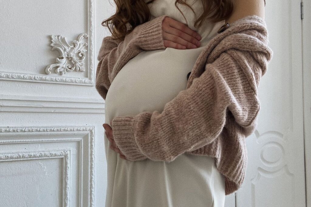 Image of a woman standing in front of a white ornate wall paneling, cradling their visible pregnant belly with both hands. The individual is wearing a white dress and an oversized knitted cardigan, with the focus on the belly suggesting the anticipation of motherhood. Image used for the article Two Cardigans for a Chic Look.