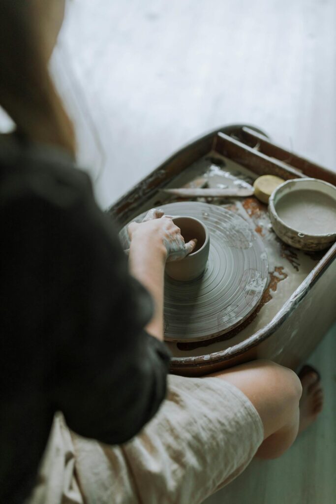 Close-up image of a woman working on a pottery wheel. Their hands are shaping a piece of clay on the spinning wheel, and there are tools and a bowl of slip (liquid clay) nearby. The focus is on the pottery process, with soft lighting and a calm, natural atmosphere. The person is sitting at the wheel, with their legs visible, and appears to be engaged in the act of molding the clay. Image used for the article outdoor hobbies for moms.