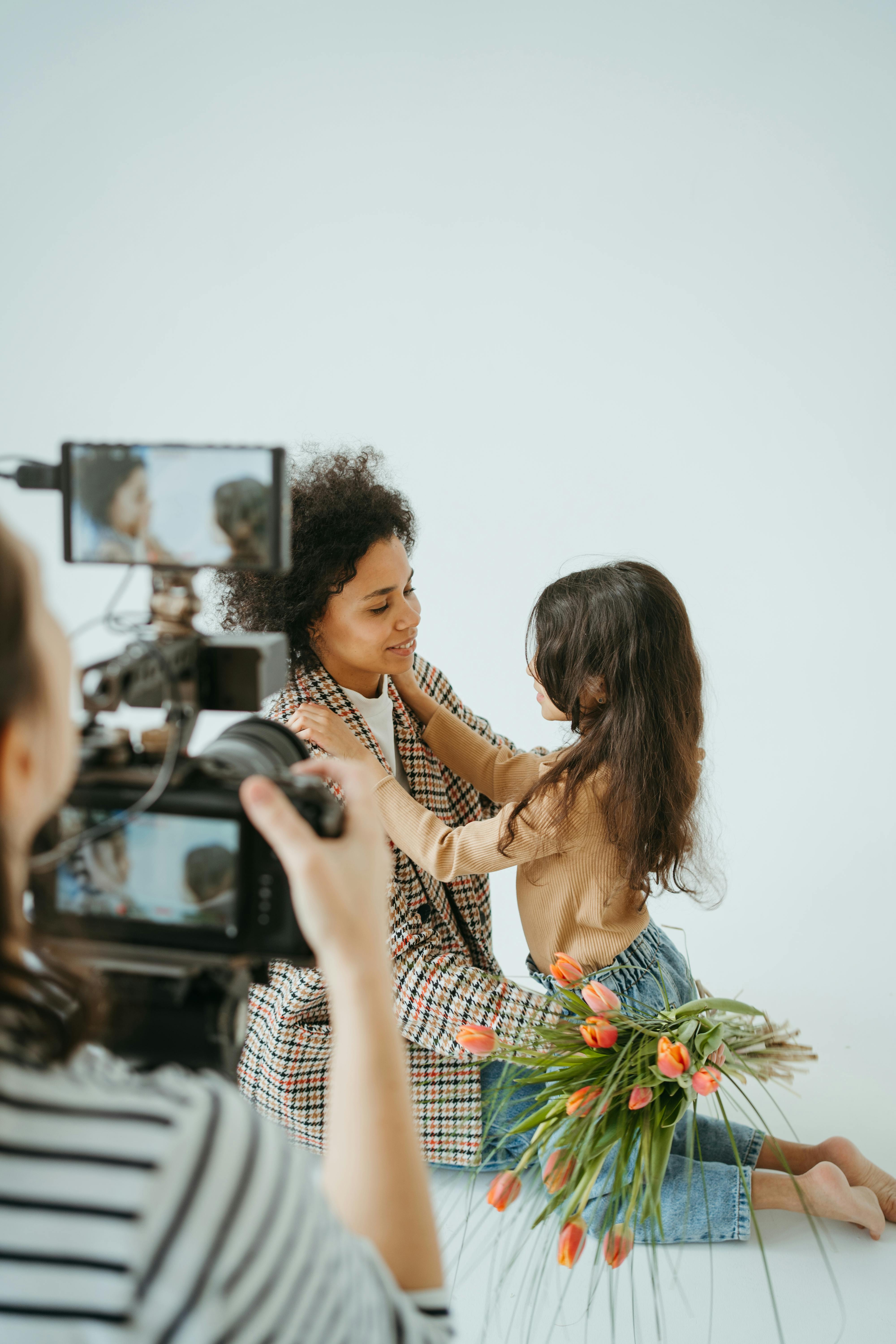 Close-up image shows of a behind-the-scenes shot of a mom and girl, used for the article spring family photo outfits. A woman with curly hair wearing a checkered coat is sitting on the floor, smiling at a young girl who is facing her. The girl, with long dark hair, is holding onto the woman's shoulders, and a bouquet of tulips is placed beside them. The scene is being recorded by someone operating a camera, partially visible in the foreground. The overall atmosphere seems warm and affectionate, capturing a tender moment between the woman and the girl.