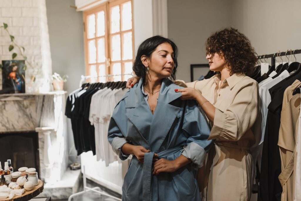 Image of wo women in a stylish boutique, with one wearing a blue trench coat while the other adjusts it for her. The woman helping is dressed in a beige outfit, carefully checking the fit. Behind them, racks of neatly arranged black and white clothes are displayed, and a well-lit room with large windows creates a warm, inviting ambiance. The scene reflects a personalized shopping experience in a chic, curated environment.