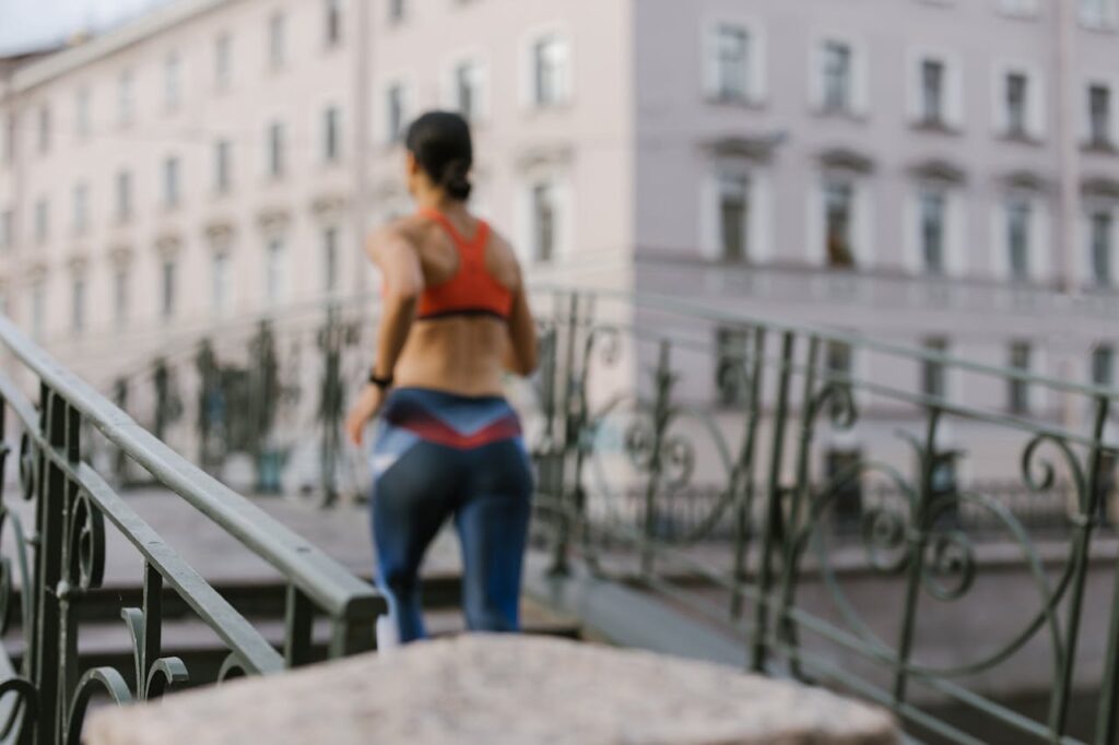 Image of a women running up a set of outdoor stairs, likely part of a bridge or a raised walkway. The runner is wearing athletic clothing, including a sports bra and leggings, and has their hair tied back. The background features an urban setting with a pastel-colored building and ornate iron railings on either side of the stairs. The image is slightly blurred, giving a sense of motion and activity as the runner moves upward. Showing Creative & Craft Hobbies for Empty Nest Moms.