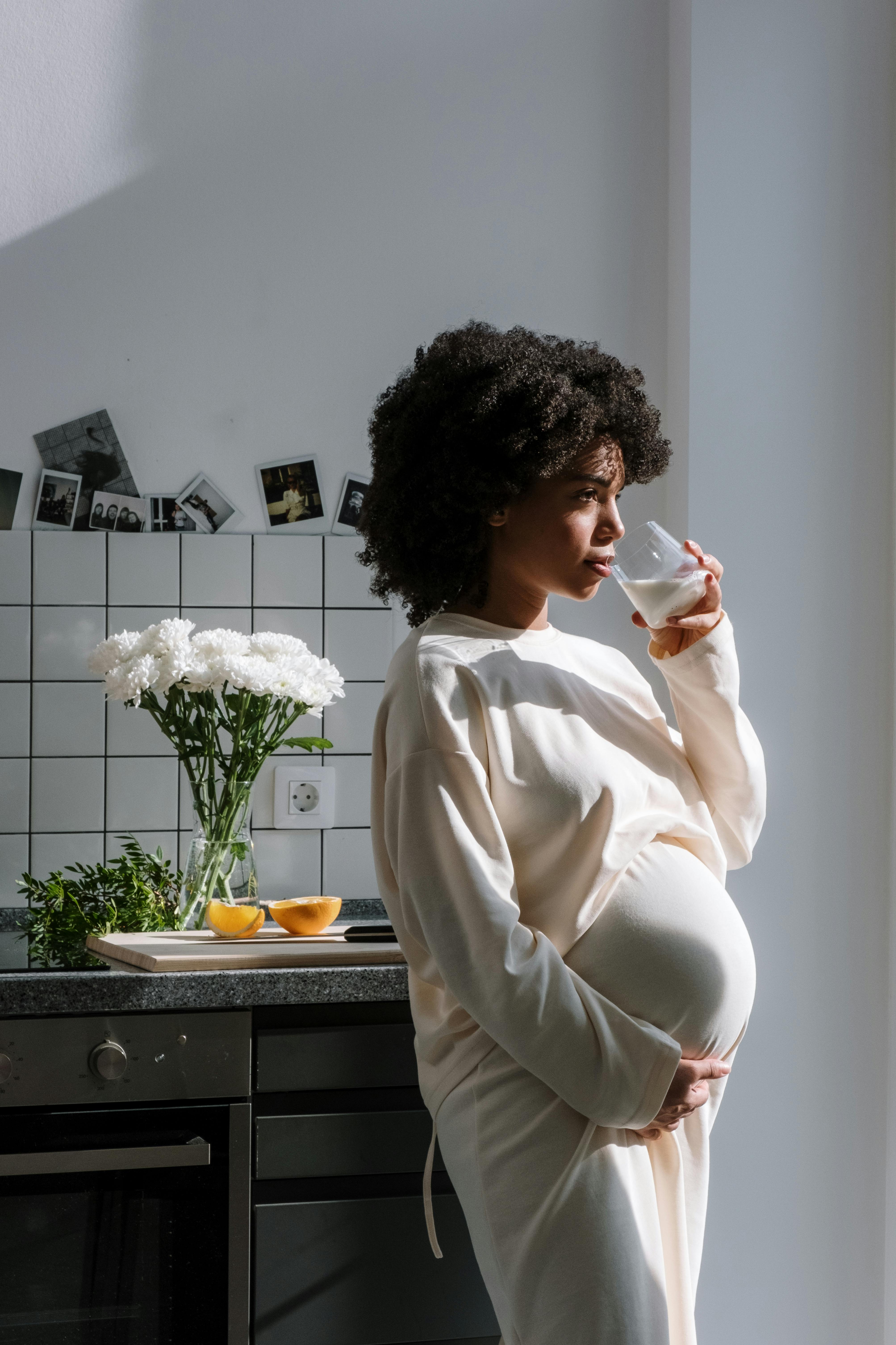 Image of a pregnant woman stands in a kitchen, holding a mug, with sunlight streaming in from the side. The individual is wearing a white long-sleeve dress and is facing away from the camera, looking out of frame. The kitchen features white tiles, and there are white flowers in a vase on the counter alongside two oranges and a bowl of eggs. There are photos attached to the wall above the counter. Image used for the article Pregnancy Capsule Wardrobe.