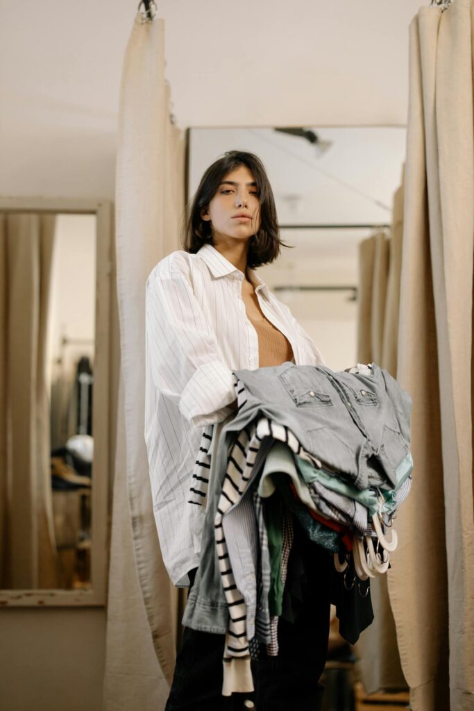 Image of a young woman standing in a fitting room holding a large pile of folded clothes. She is dressed in a loose, oversized white button-up shirt over a brown top, with a casual, effortless look. Her expression is calm and slightly pensive, and behind her are light-colored curtains separating the changing areas. The clothes in her arms include a mix of striped and solid garments, along with a pair of denim shorts on top. The setting appears to be a relaxed regular wardrobe updates.