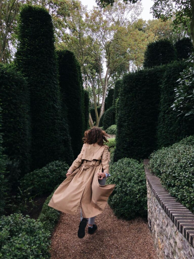 A person with curly hair is running along a narrow gravel path through a garden maze of tall, meticulously trimmed hedges. They are wearing a beige trench coat that flares behind them as they move. The setting is serene, surrounded by lush greenery with trees towering above the maze. Image used for the article Hobbies for Empty Nest Moms.