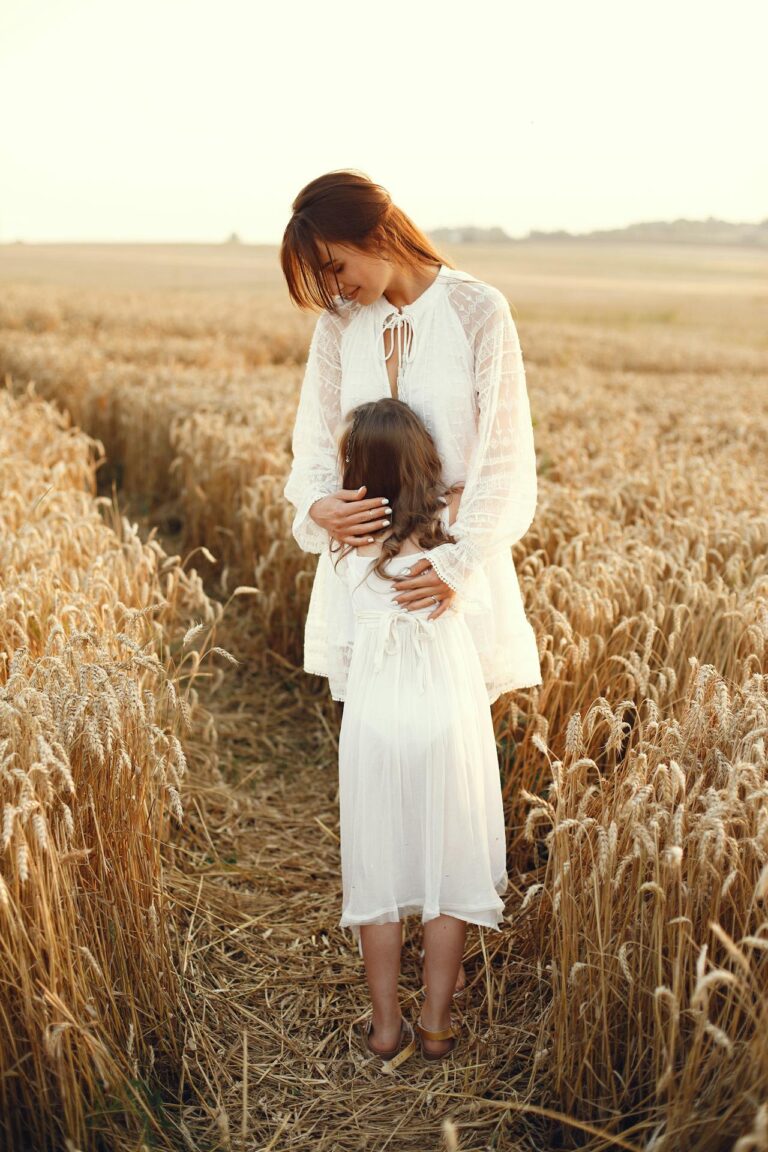 Image of a woman and a young girl are standing in a golden wheat field, embraced closely. Both are dressed in flowing white dresses, with the woman's arms gently wrapped around the girl. The scene is warm, peaceful, and sunlit, evoking feelings of love and tenderness. The wheat field stretches into the horizon, creating a serene and natural backdrop. Image used for the article Single Mom Family Picture Ideas.