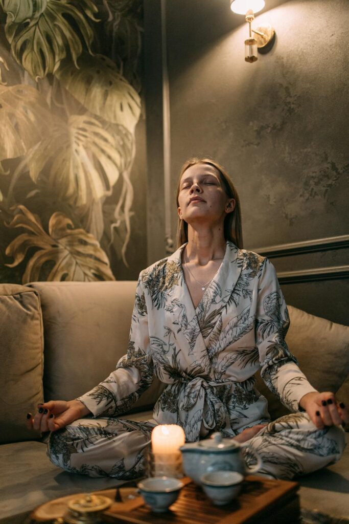 Close-up image of a woman sitting in a meditative pose on a comfortable couch. She is dressed in light-colored, leaf-patterned loungewear with her eyes closed, exuding a sense of calm and relaxation. A soft light from a wall-mounted lamp above her illuminates the serene environment. The background features botanical-themed wall decor, adding to the tranquil ambiance. A lit candle and tea set are placed in front of her, enhancing the cozy, peaceful mood.