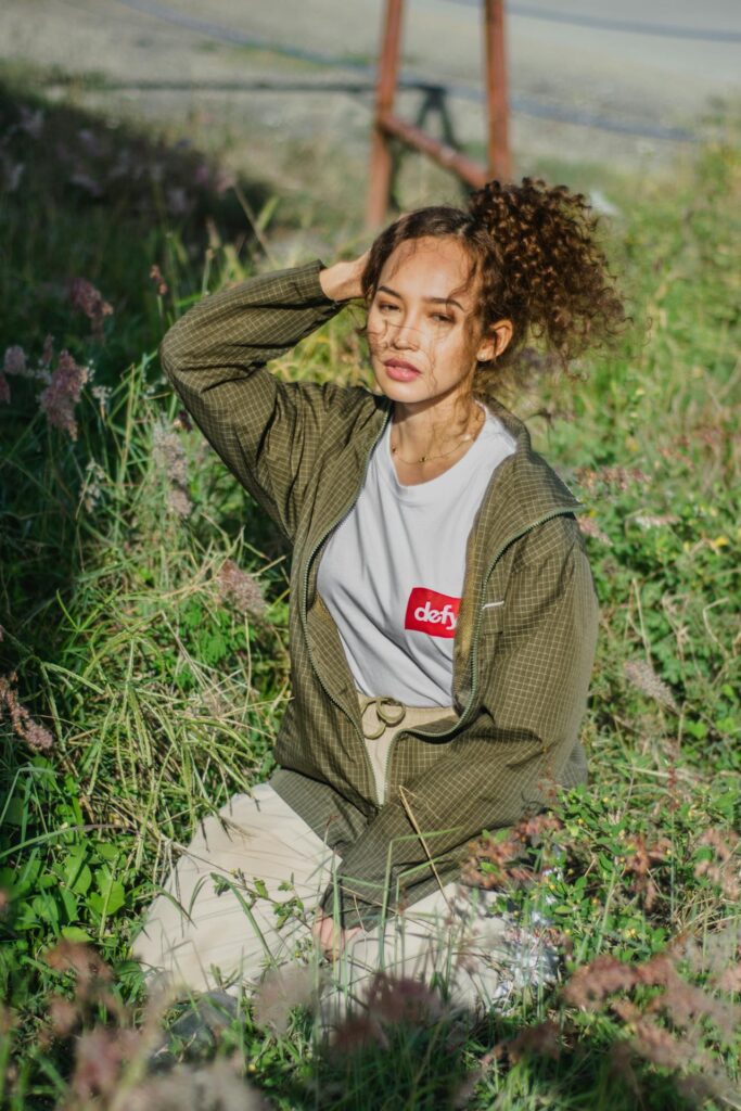 Image of a young woman sitting in a field of tall grass, wearing a casual outdoor outfit. She has on a green zip-up jacket over a white t-shirt with a red graphic that says 'defy.' Her light beige pants are cinched at the waist with a belt, and her curly hair is tied up in a loose ponytail. She holds one hand to her head, and sunlight casts soft shadows across her face. Image used for the article Don’t Forget Styling Your Hair.