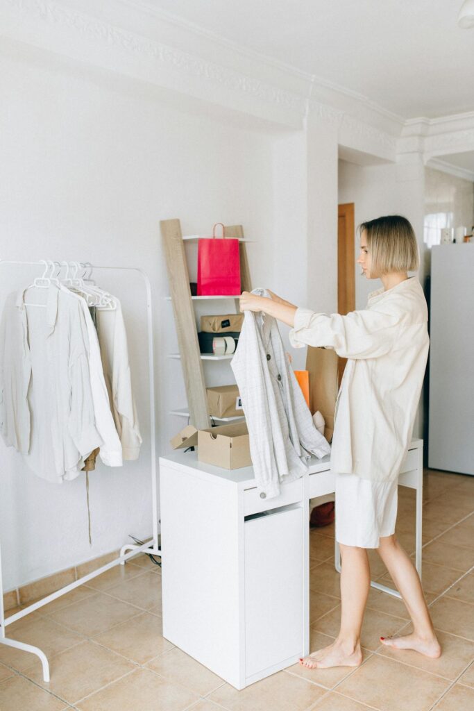 A woman is standing barefoot in a minimalist room, holding a light-colored garment while inspecting it. She is dressed in a casual cream-colored outfit, matching the soft tones of the room. Nearby, a clothes rack displays a few light garments, and a shelf behind her holds a pink shopping bag and various boxes. The overall scene exudes simplicity and organization, with a focus on fashion and a cozy atmosphere. Image used for the article how to improve your fashion style