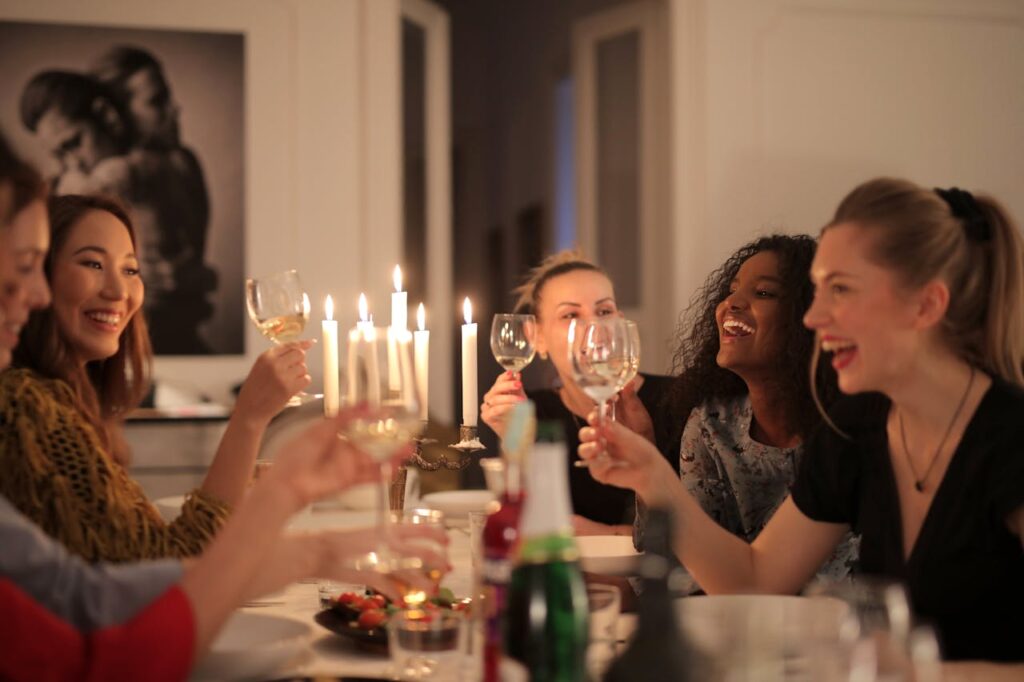 Close-up image of a group of five women sitting around a dinner table, enjoying each other's company. They are all smiling and raising glasses in a toast. The table is set with a few bottles, food, and lit candles, creating a warm and intimate atmosphere. In the background, there is a black-and-white portrait on the wall. The overall mood is joyful and celebratory, suggesting a gathering or party among friends. Image used for the article stay-at-home mom capsule wardrobe