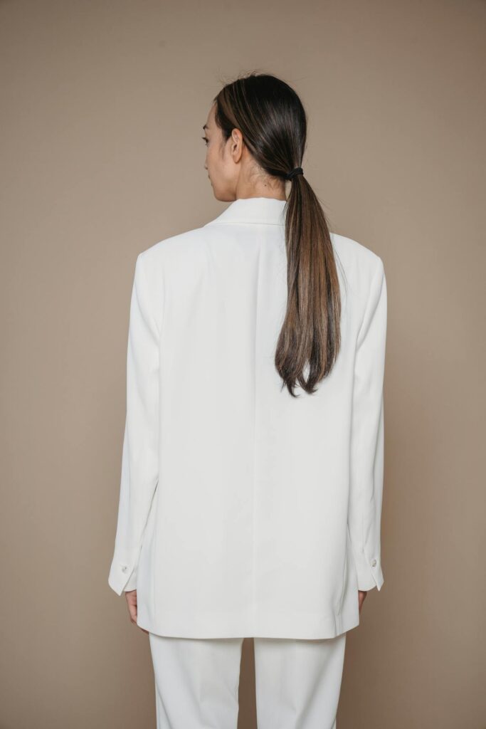 Image of a woman standing with their back to the camera, wearing a white blazer and white pants. The person has long hair tied in a low ponytail. The background is a solid light brown color.  Representing Fashion colors for Moms in Their 30s.