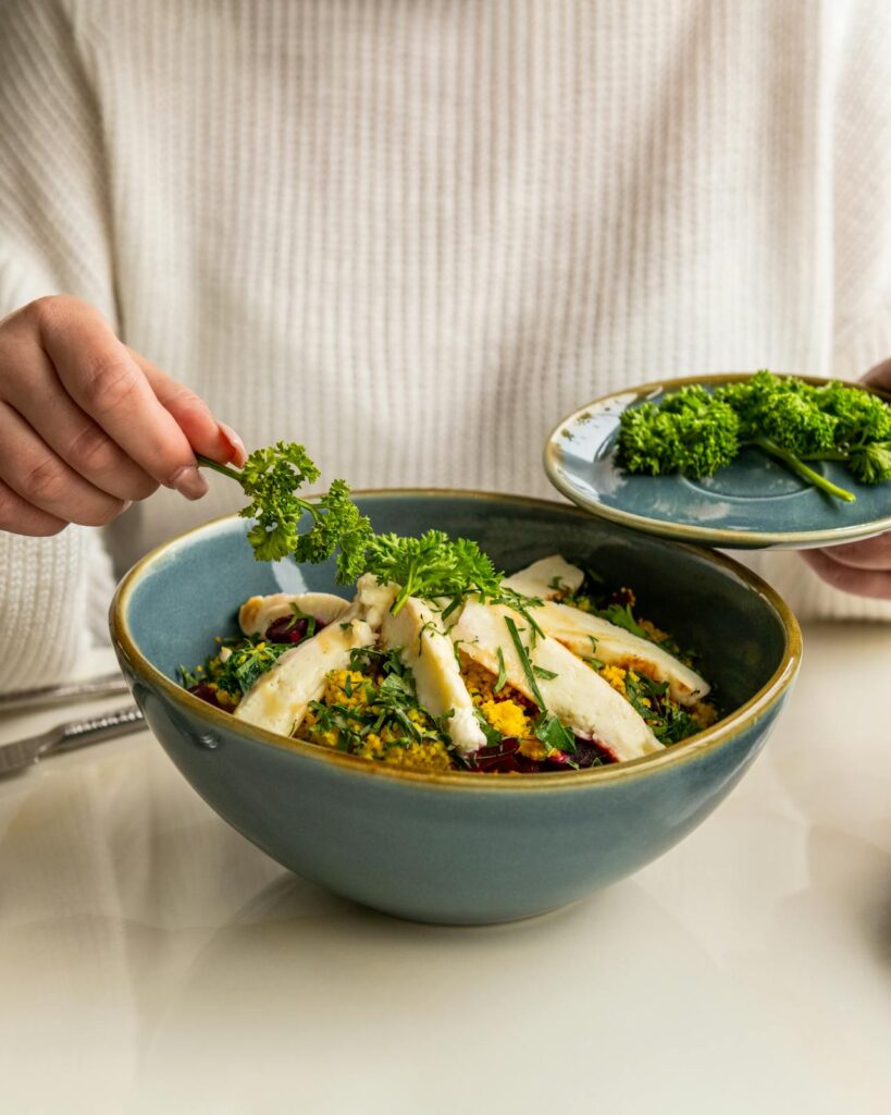 Image of a person adding fresh parsley to a dish in a bowl. The dish appears to be a vibrant salad or grain bowl, topped with sliced cheese or chicken, herbs, and possibly vegetables. The bowl is a large, rustic ceramic with a blue-green glaze. The person is wearing a white sweater, and they are carefully garnishing the dish by hand while holding a small plate of additional parsley. The setting is clean and minimal, with a white table surface beneath the bowl.