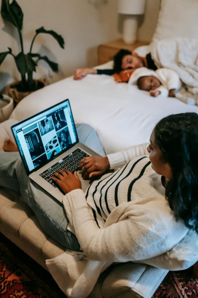 Close-up image of a woman sitting on a bed with a laptop on their lap watching a video. In the background, another person is lying down, partially covered by bedding. The room feels cozy with a plant on the left and pillows scattered around. Representing mom fashion 2024/2025.