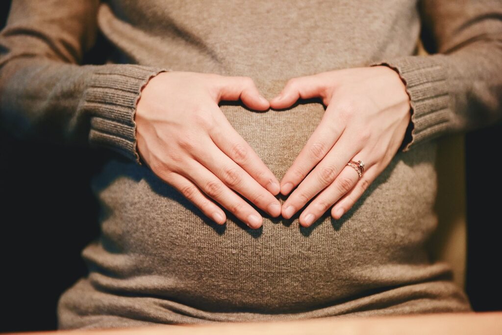 A close-up image of a woman wearing a grey sweater forming a heart shape with their hands over their stomach. The person is wearing a ring on the ring finger of their left hand, suggesting they might be engaged or married. The focus on the hands and the heart shape could imply love, affection, or could be related to pregnancy. Image used for the article An Oversized Sweatshirt.