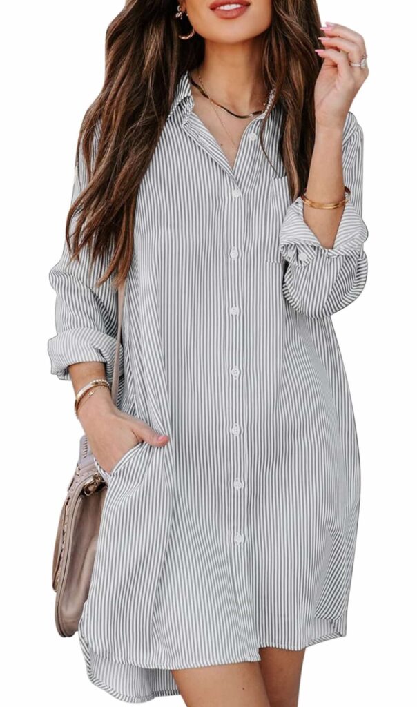  An image of a woman wearing a long-sleeved, knee-length shirt dress with vertical stripes. The dress features a collar, button-down front, and appears to be cinched at the wrists. The person is also carrying a light-colored shoulder bag and is posing with one hand touching their hair.