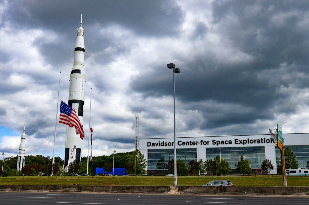 Image of a large rocket stands tall outside the Davidson Center for Space Exploration, with the American flag prominently displayed at half-mast beside it. The sky is cloudy, casting a dramatic atmosphere over the scene. The building, modern and expansive, serves as a backdrop, with large glass windows and the center's name clearly visible in bold black letters. Trees and a few cars can be seen in the foreground, and a road runs along the bottom of the image. The overall scene highlights a space exploration facility with a patriotic and scientific focus.