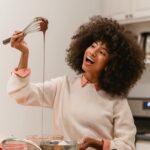 Image of a cheerful woman with curly hair is holding a whisk with melted chocolate dripping from it, smiling as she cooks in a kitchen. She is dressed in a light-colored sweater with a pink collar and is surrounded by various cooking utensils on the countertop. Image used for the article Hobbies for Ladies.