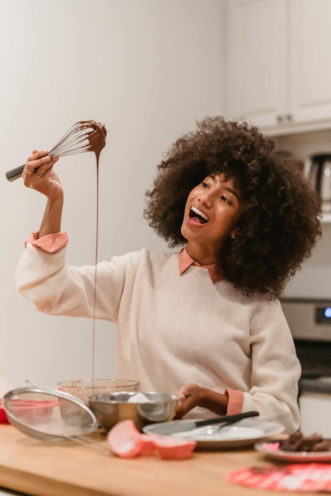 Image of a cheerful woman with curly hair is holding a whisk with melted chocolate dripping from it, smiling as she cooks in a kitchen. She is dressed in a light-colored sweater with a pink collar and is surrounded by various cooking utensils on the countertop. Image used for the article Hobbies for Ladies.