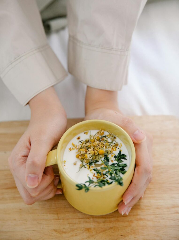 A close-up image of a person holding a yellow mug with both hands. The mug is filled with a creamy beverage, topped with dried chamomile flowers and fresh thyme sprigs. The person is wearing a light-colored, long-sleeved shirt, and the scene is set on a wooden surface, creating a cozy and calming atmosphere.