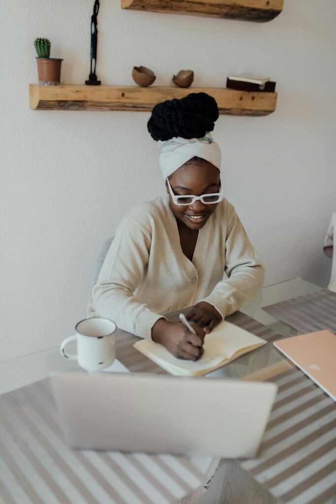 Image of a woman is sitting at a table, writing in a notebook while smiling. She is wearing white-framed glasses, a light beige cardigan, and has her hair wrapped in a headscarf. In front of her is an open laptop, and a mug sits to her left. The background features wooden shelves mounted on the wall, holding small potted plants and decorative items, creating a cozy and organized workspace.