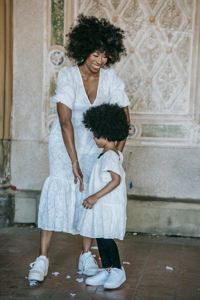 Image of a woman and a child, both wearing white dresses, are sharing a tender moment. The woman is smiling and gently holding the child, who stands in front of her. They both have curly, voluminous hair, and the child is looking down while leaning towards the woman. They are wearing matching white sneakers. The background features an ornate wall with intricate patterns, adding a decorative, vintage feel to the setting. The atmosphere is warm and joyful, highlighting a beautiful connection between the two.