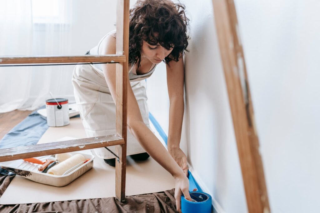 Image of a woman preparing to paint a room by carefully applying blue painter's tape along the base of a white wall. They are kneeling on the floor, wearing a light-colored apron. Nearby, there is painting equipment, including a paint roller tray, a roller, and a can of white paint. The person is working in a well-lit room, possibly near a window, with a ladder visible in the foreground. The floor is protected with a brown and blue covering to prevent paint from spilling. The scene conveys a focused, methodical approach to interior painting. Image used for the article female dominated hobbies.