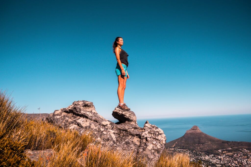 Image of a woman stands on a rocky outcrop overlooking a vast landscape with clear blue skies. She is wearing a black tank top, teal shorts, and hiking shoes, gazing into the distance. The scenery includes grassy terrain, distant mountains, and a view of the ocean or large body of water in the background representing hobbies for ladies over 50.