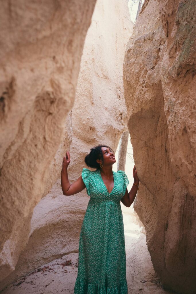 A woman stands in a narrow, sandy canyon between tall, light-colored rock formations. She is wearing a long, flowing green dress with short sleeves, featuring a subtle floral pattern. Her hands are gently touching the canyon walls on either side as she looks up thoughtfully. The natural light softly illuminates her face and the surrounding rocks, creating a serene and tranquil atmosphere. The scene suggests a peaceful moment of exploration in a unique natural landscape. Showing Choosing Outfits for Your Fall Family Photoshoot.