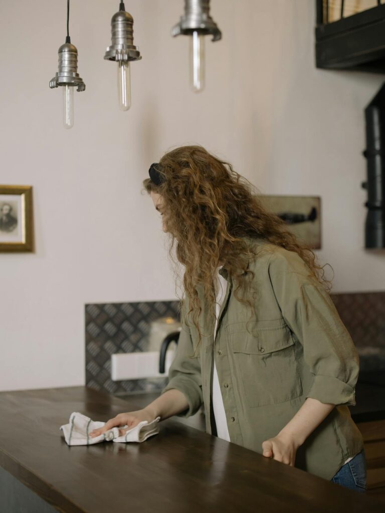 Image of a woman with long, curly hair is cleaning a wooden countertop in a cozy, rustic kitchen. She is wiping the surface with a cloth while wearing an olive-green button-up shirt and a black headband. The kitchen has a warm, industrial style, featuring hanging metal pendant lights above the counter and a simple backsplash with dark tones. A kettle is visible in the background, along with framed wall art that adds a personal touch to the space. The overall ambiance is quiet and homely, with the woman focused on her task.