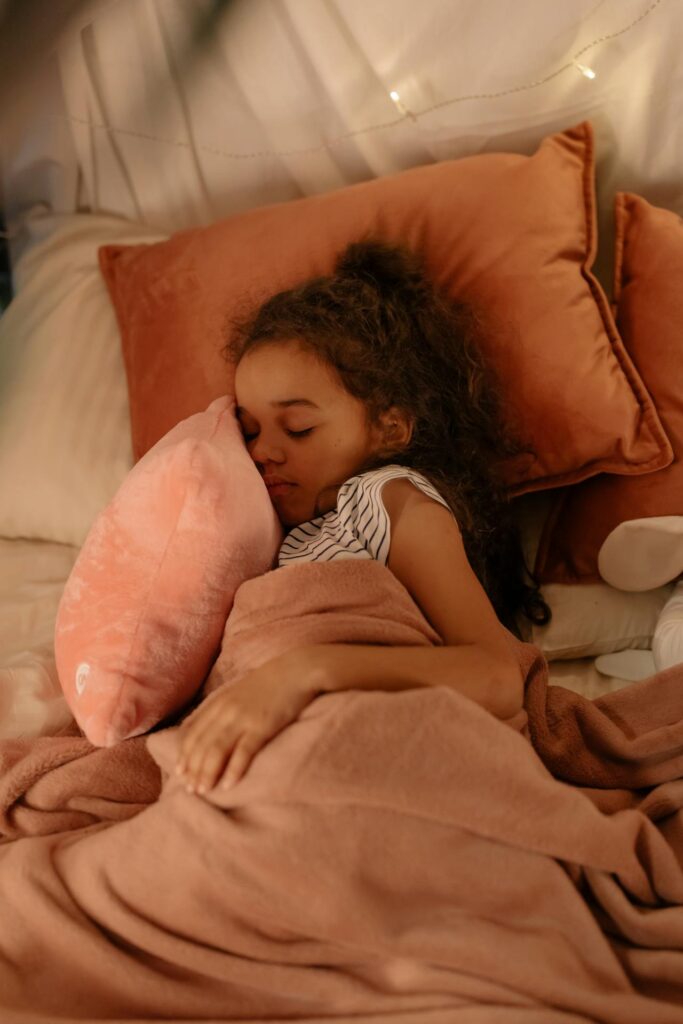 Image of a young girl peacefully asleep in bed, cuddling a soft pink pillow. She is wrapped in a cozy blanket of a similar pink hue, adding warmth and comfort to the scene. The bed is adorned with large, plush pillows in shades of coral and peach, enhancing the snug atmosphere. Gentle, ambient lighting and soft bedding create a calming and serene environment, perfect for a restful sleep. The girl's relaxed expression and cozy surroundings make the scene feel especially tranquil and comforting.