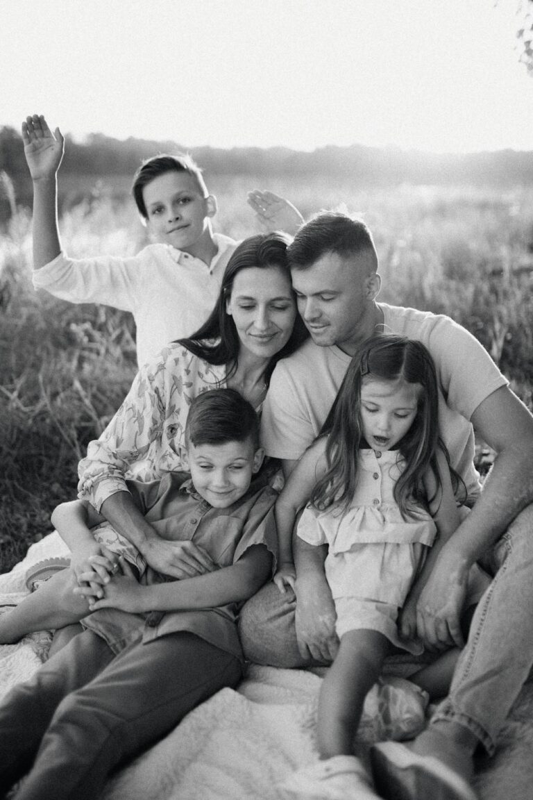 A black and white photo of a family sitting together in a peaceful outdoor setting. The parents are sitting in the center, with the father gently leaning toward the mother, their faces close and serene. Two young children, a boy and a girl, are sitting in front of them, looking down. Another older boy stands behind them, raising one hand playfully. The family is sitting on a blanket, surrounded by soft, natural light, giving the image a warm and affectionate feel. The background appears to be a grassy field, adding to the relaxed atmosphere. Image used for the article Family Photo Dresses for Mom.