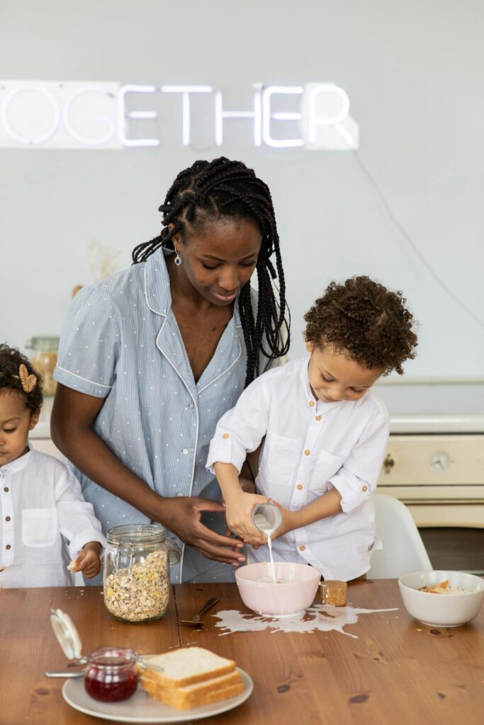 Image of a woman in pajamas is helping two children prepare breakfast at a wooden table. The boy is pouring milk into a bowl of cereal, spilling some on the table, while the woman guides him. The girl on the left, dressed similarly in a white shirt, watches. The background shows a simple kitchen, and a neon sign above them reads "TOGETHER." The scene captures a warm, familial moment of bonding over breakfast. Image used for the article A Simple Stay at Home Mom Schedule.