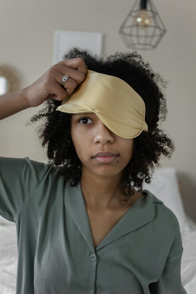 Close-up image of a woman with curly hair is adjusting a yellow sleep mask on her forehead, with one side covering her eye. She is wearing a sage green button-up pajama top and has a neutral expression on her face. The room in the background is softly lit, with a modern geometric light fixture hanging from the ceiling and a bed behind her, adding to the cozy and relaxed atmosphere. A small ring is visible on her hand as she lifts the mask. Showing how to wake up at 5am.