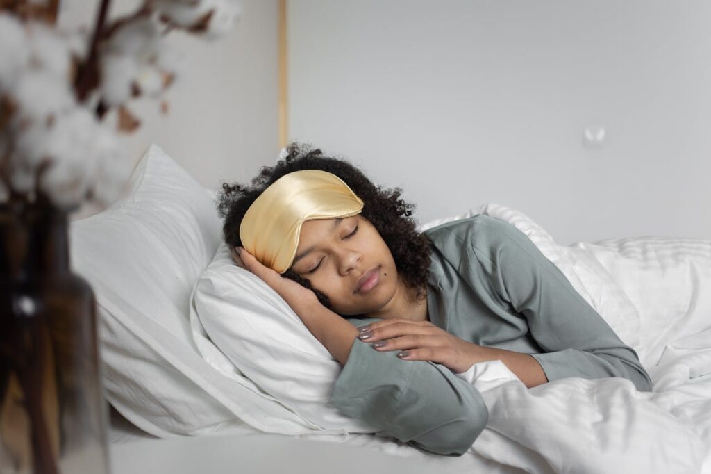 Image of a woman is peacefully sleeping in bed, wearing a satin sleep mask over her eyes. She is resting her head on a white pillow, with her hand gently placed by her face. Her curly hair peeks out from under the mask, and she is dressed in a soft, long-sleeved gray shirt. The bedding is white, and the room appears calm and serene, with a blurred plant visible in the foreground on the left side of the image.